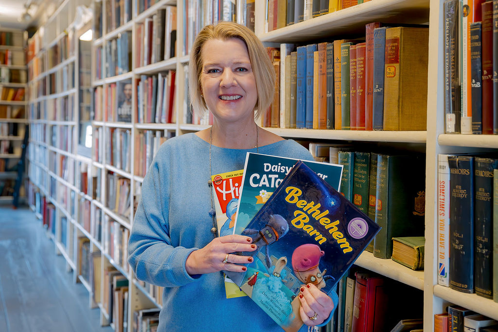 Debra Westgate-Silva: Standing by bookshelf holding three books.