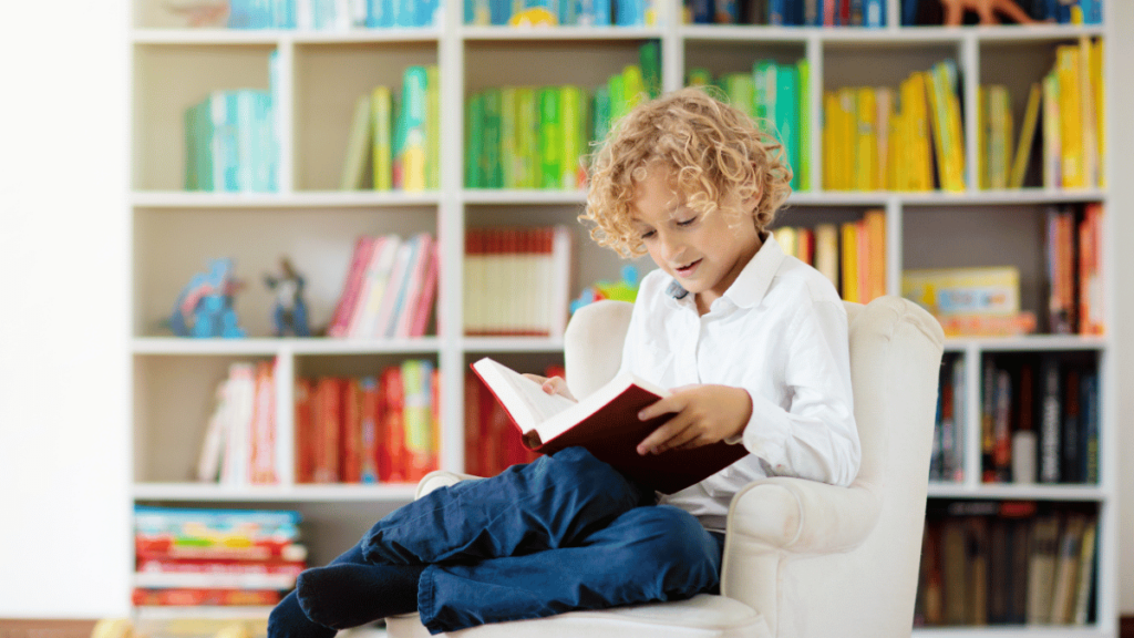 Young child reading a book while sitting in an armchair. Colorful bookshelf behind him.