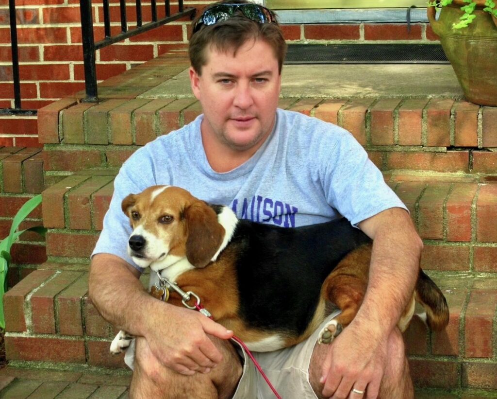 Frank Payne author Headshot (sitting on brick stairs), holding beagle.